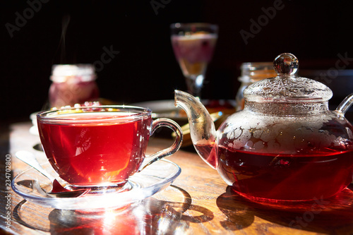 From a clear glass teapot, pour hot tea into a transparent cup. against sunlight. warm atmosphere. There are many different desserts in the background. the whole composition on the wooden table