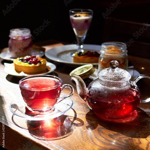 From a clear glass teapot, pour hot tea into a transparent cup. against sunlight. warm atmosphere. There are many different desserts in the background. the whole composition on the wooden table