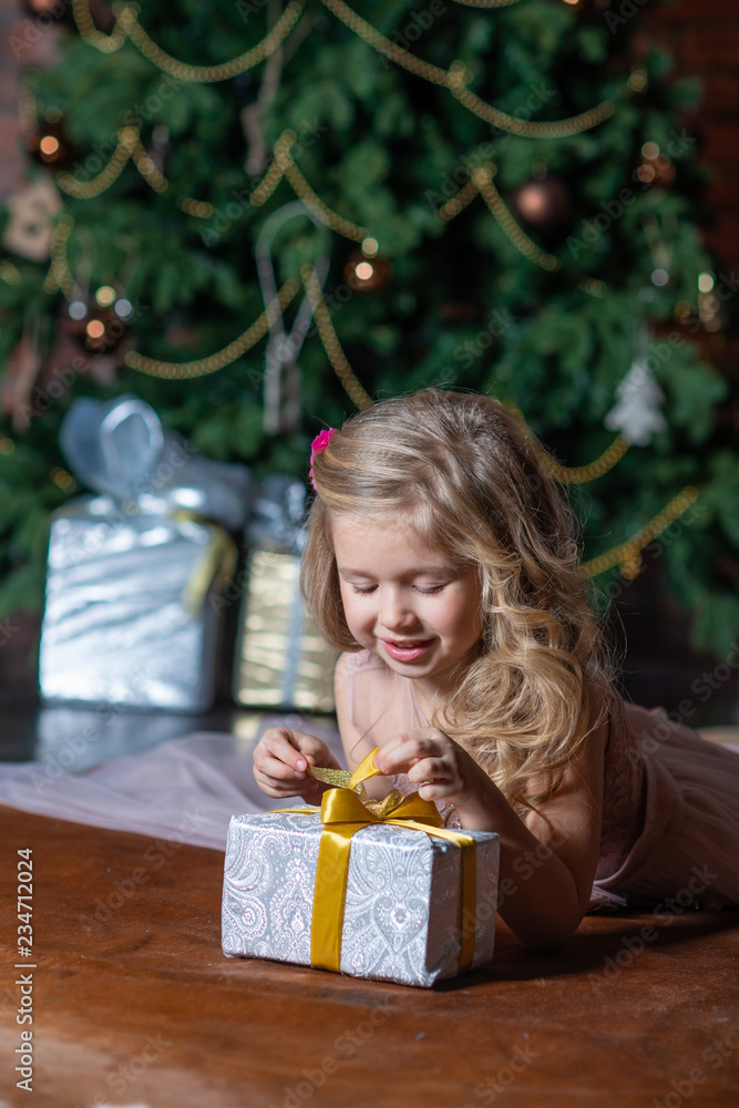 Little smiling girl opens a gift box on the floor next to the Christmas tree Little smiling girl opens a gift box on the floor next to the Christmas tree