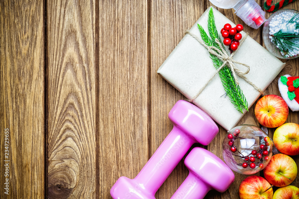 Fitness, healthy and active lifestyles Christmas gift concept, dumbbells, apples, gift box with Christmas ornaments decoration on wood background. Top view with copy space. Fitness, healthy and active lifestyles Christmas gift concept, dumbbells, apples, gift box with Christmas ornaments decoration on wood background. Top view with copy space.