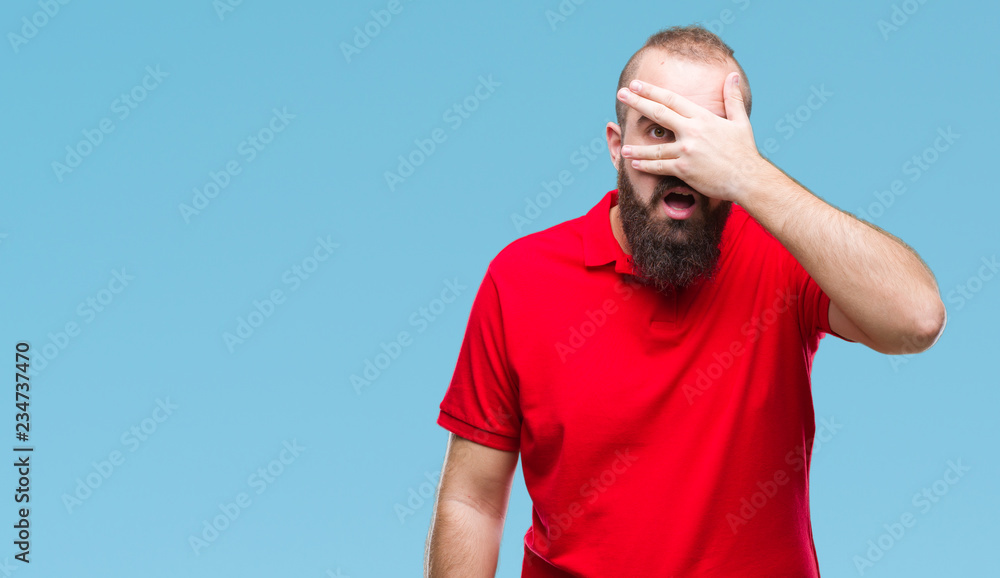 Young caucasian hipster man wearing red shirt over isolated background peeking in shock covering face and eyes with hand, looking through fingers with embarrassed expression. Young caucasian hipster man wearing red shirt over isolated background peeking in shock covering face and eyes with hand, looking through fingers with embarrassed expression.
