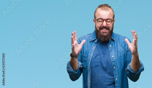 Young caucasian hipster man wearing glasses over isolated background crazy and mad shouting and yelling with aggressive expression and arms raised. Frustration concept.