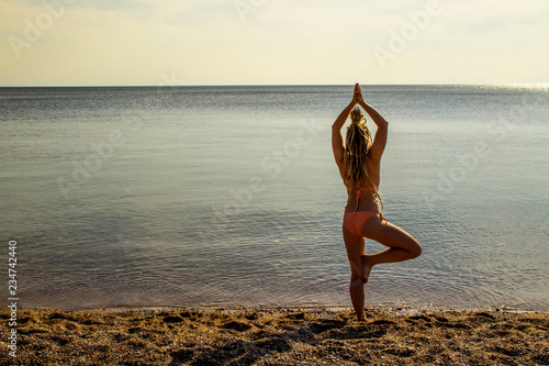 Attractive fit young modern woman in vibrant orange bikini do yoga tree pose  vrksasana  by the sea on sunny sandy beach  calm waters  warm colors. Inner balance concept.