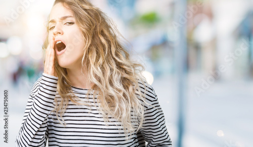 Beautiful young blonde woman wearing stripes sweater over isolated background bored yawning tired covering mouth with hand. Restless and sleepiness.