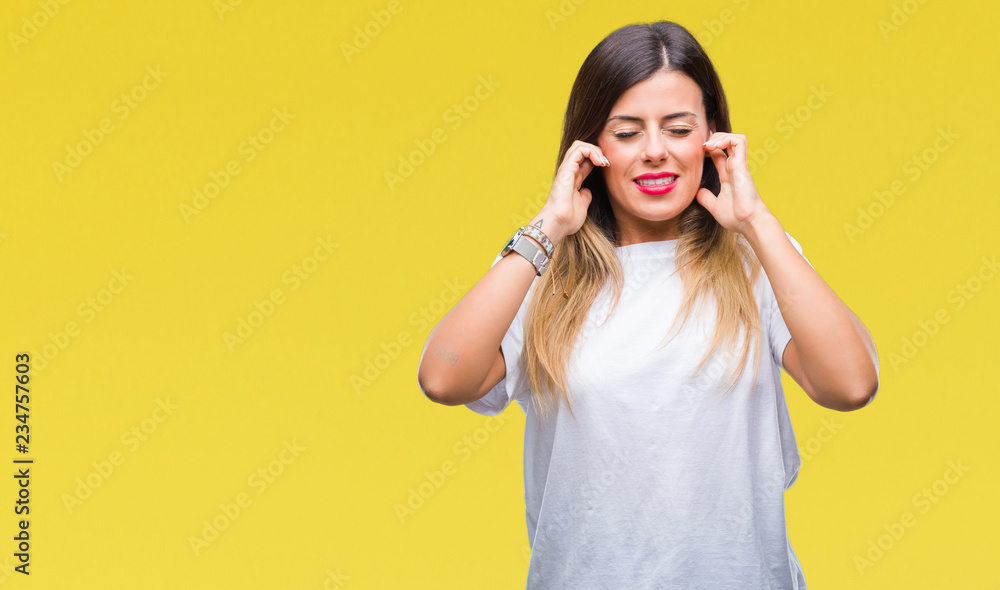 Young beautiful woman casual white t-shirt over isolated background covering ears with fingers with annoyed expression for the noise of loud music. Deaf concept. Young beautiful woman casual white t-shirt over isolated background covering ears with fingers with annoyed expression for the noise of loud music. Deaf concept.