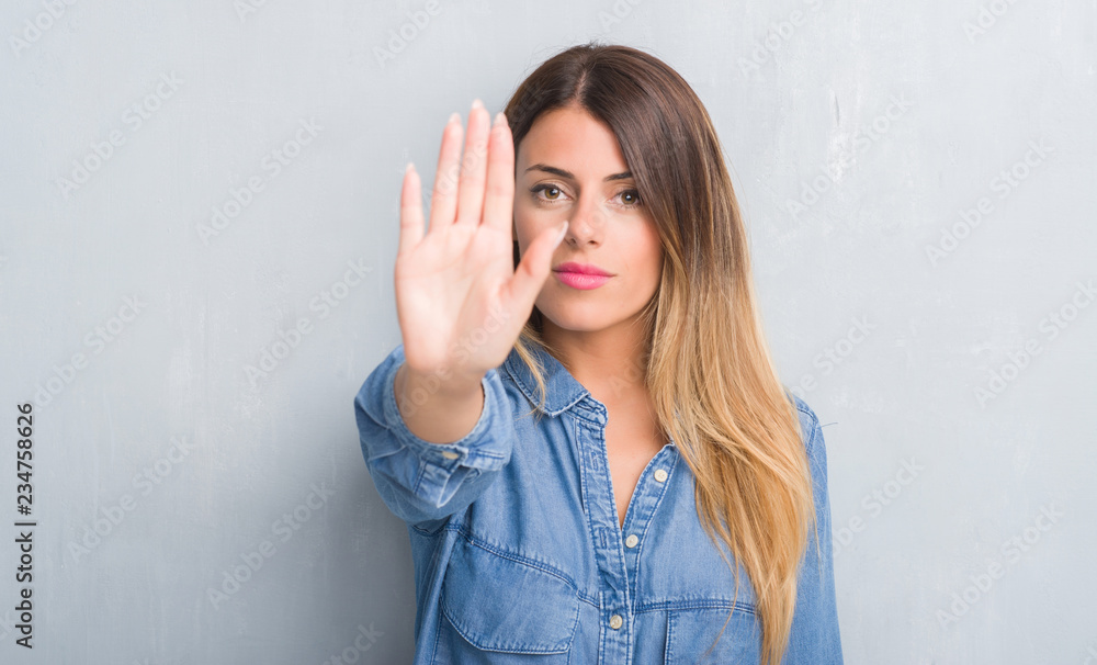 Young adult woman over grey grunge wall wearing denim outfit with open hand doing stop sign with serious and confident expression, defense gesture Young adult woman over grey grunge wall wearing denim outfit with open hand doing stop sign with serious and confident expression, defense gesture
