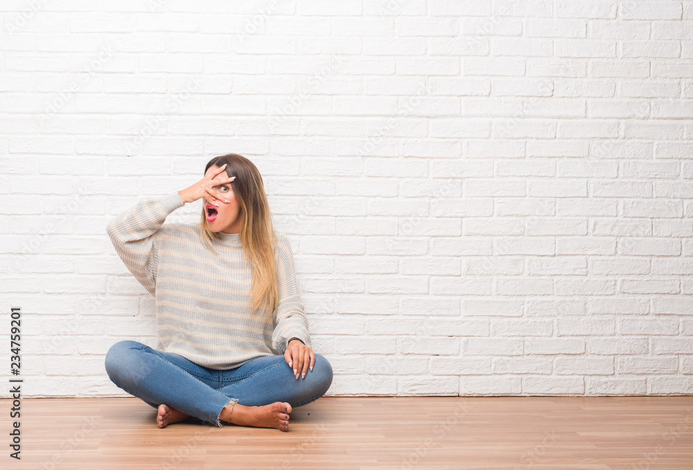 Young adult woman sitting on the floor over white brick wall at home peeking in shock covering face and eyes with hand, looking through fingers with embarrassed expression. Young adult woman sitting on the floor over white brick wall at home peeking in shock covering face and eyes with hand, looking through fingers with embarrassed expression.