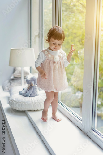 Little cute sweet smiling girl in pastel pink dress standing on the window sill in bright light living room at home and smiling. Childhood, preschool, youth, relax concept