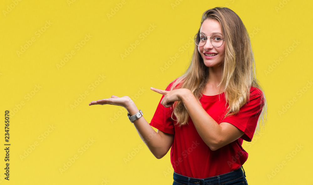 Young beautiful blonde woman wearing glasses over isolated background amazed and smiling to the camera while presenting with hand and pointing with finger. Young beautiful blonde woman wearing glasses over isolated background amazed and smiling to the camera while presenting with hand and pointing with finger.