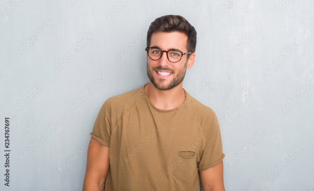 Handsome young man over grey grunge wall wearing glasses looking away to side with smile on face, natural expression. Laughing confident. - obrazy, fototapety, plakaty 