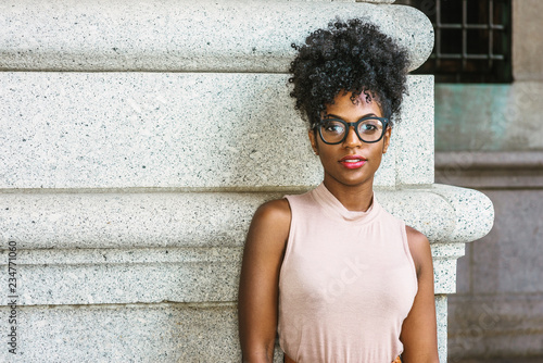 Obraz na płótnie Portrait of Young African American Woman in New York. Young black female college student with afro hairstyle wearing sleeveless light color top, eye glasses, standing by column on street on campus.. w salonie