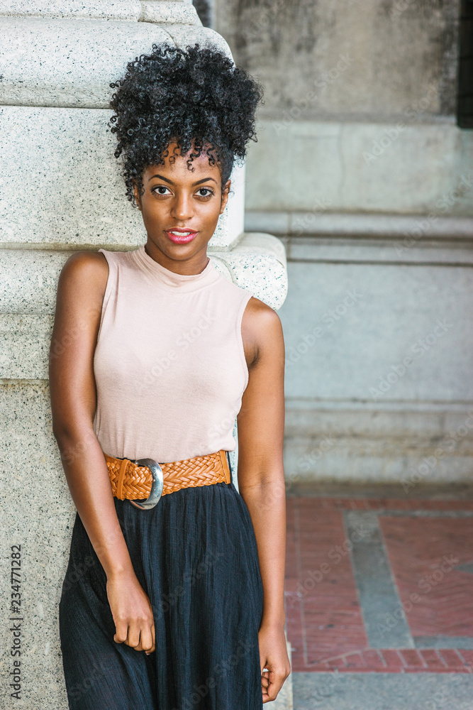Portrait of Young African American Woman in New York. Young black female  college student with afro hairstyle wearing sleeveless light color top,  brown belt, black skirt, standing by column on street.. Stock