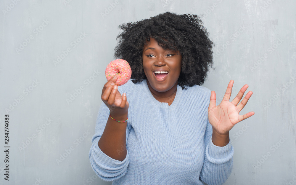 Young african american woman over grey grunge wall eating pink donut very happy and excited, winner expression celebrating victory screaming with big smile and raised hands Young african american woman over grey grunge wall eating pink donut very happy and excited, winner expression celebrating victory screaming with big smile and raised hands