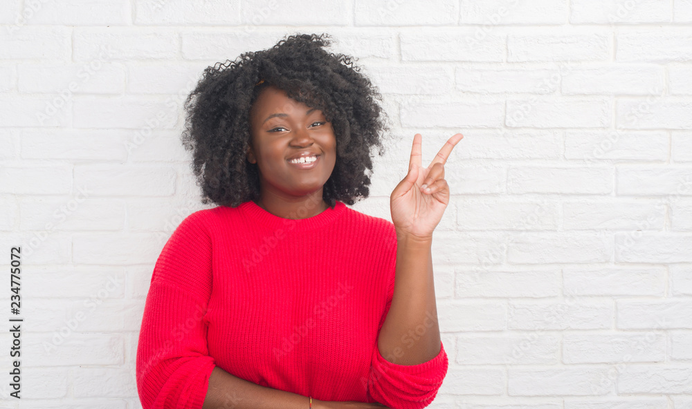 Young african american plus size woman over white brick wall smiling with happy face winking at the camera doing victory sign. Number two. Young african american plus size woman over white brick wall smiling with happy face winking at the camera doing victory sign. Number two.