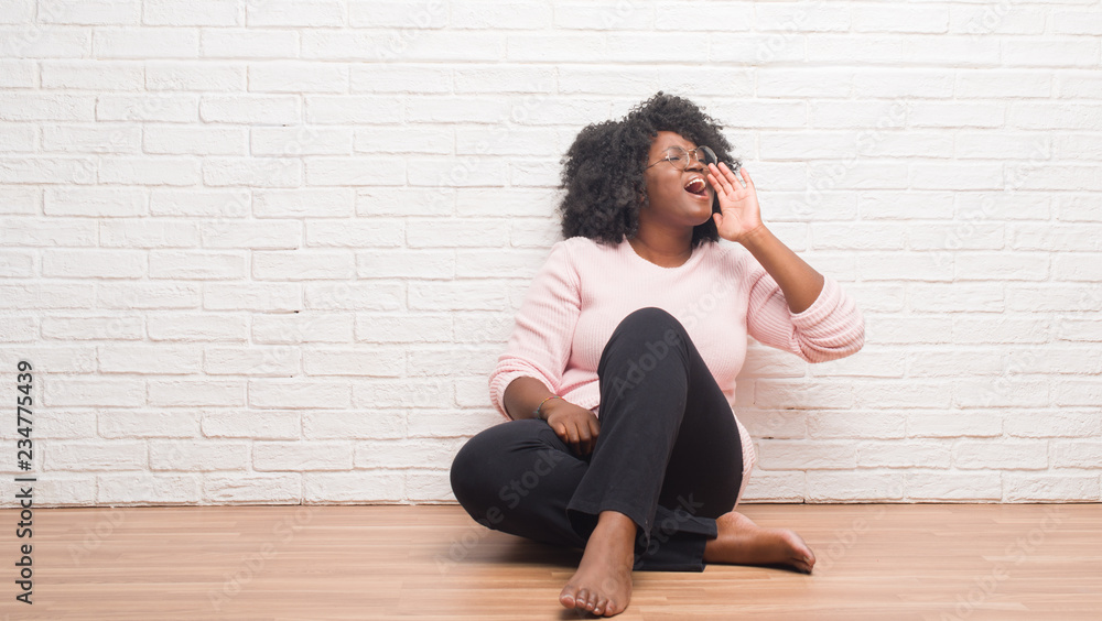 Young african american woman sitting on the floor at home shouting and screaming loud to side with hand on mouth. Communication concept. Young african american woman sitting on the floor at home shouting and screaming loud to side with hand on mouth. Communication concept.
