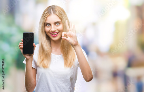 Young beautiful blonde woman showing smartphone over isolated background doing ok sign with fingers, excellent symbol