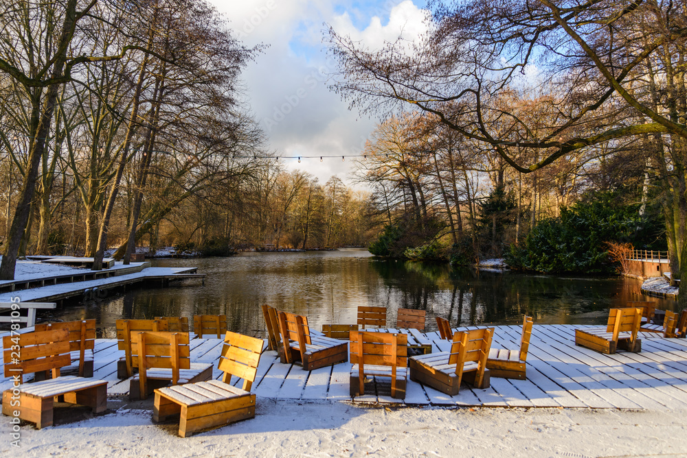 Scenery of frozen lake, snow cover wooden outdoor chairs, seats and deck waterside and forest in winter atmosphere. View from Café am Neuen See inside Tiergarten park in Berlin with sunset light.
