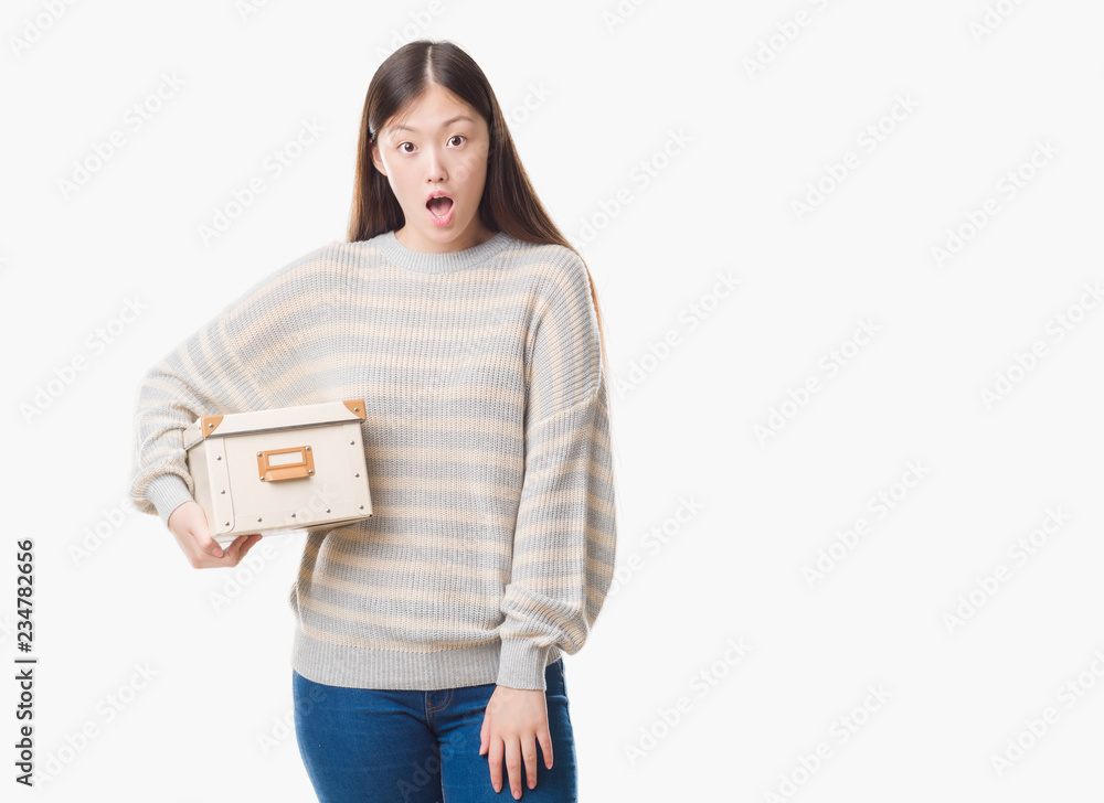 Young Chinese woman over isolated background holding a box scared in shock with a surprise face, afraid and excited with fear expression Young Chinese woman over isolated background holding a box scared in shock with a surprise face, afraid and excited with fear expression