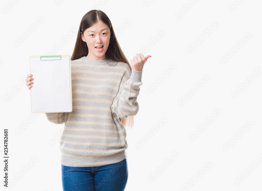 Young Chinese woman over isolated background holding clipboard pointing and showing with thumb up to the side with happy face smiling Young Chinese woman over isolated background holding clipboard pointing and showing with thumb up to the side with happy face smiling