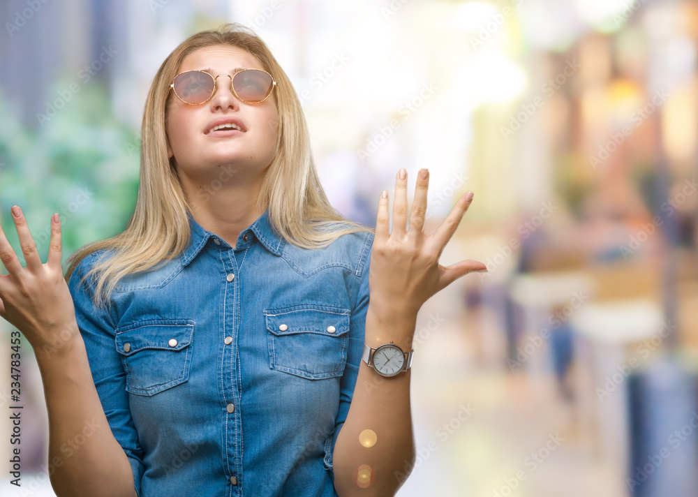 Young caucasian woman wearing sunglasses over isolated background crazy and mad shouting and yelling with aggressive expression and arms raised. Frustration concept.