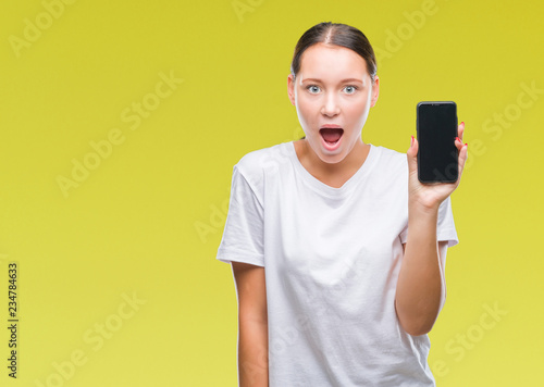 Young beautiful caucasian woman showing smartphone screen over isolated background scared in shock with a surprise face, afraid and excited with fear expression