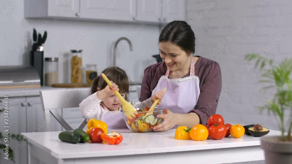 Happy little daughter with down syndrome stirring fresh salad while preparing healthy food together with cheerful mother in the kitchen. Caring mother teaching her special needs child cooking. Happy little daughter with down syndrome stirring fresh salad while preparing healthy food together with cheerful mother in the kitchen. Caring mother teaching her special needs child cooking.