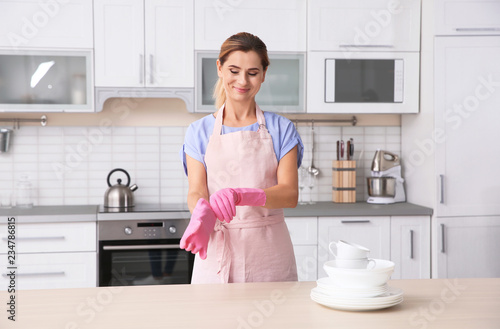 Woman wearing rubber gloves near table with clean dishes and cups in kitchen