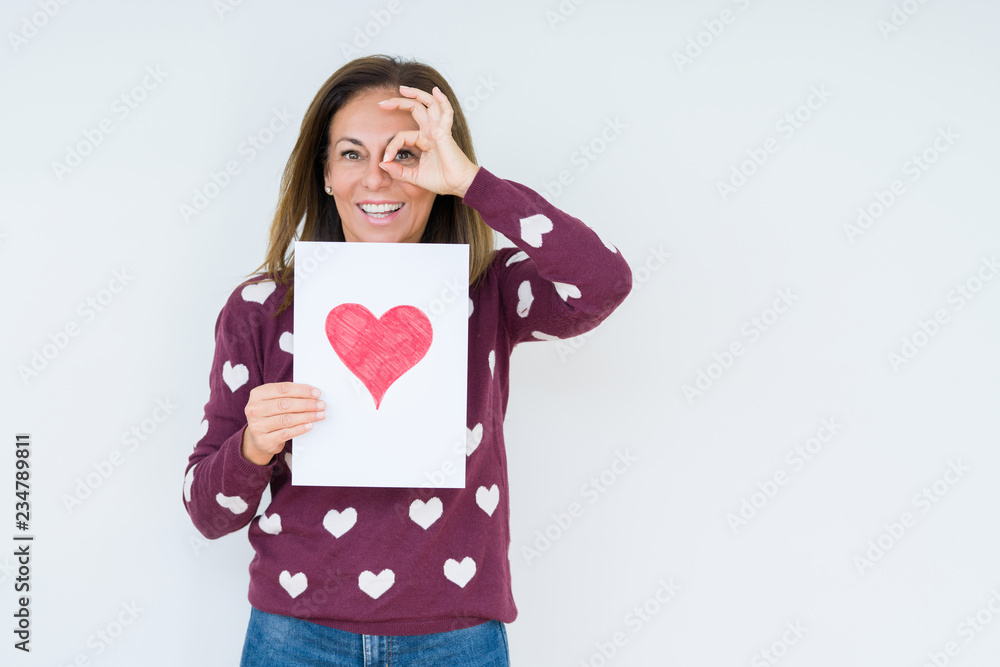 Middle age woman holding card gift with red heart over isolated background with happy face smiling doing ok sign with hand on eye looking through fingers Middle age woman holding card gift with red heart over isolated background with happy face smiling doing ok sign with hand on eye looking through fingers