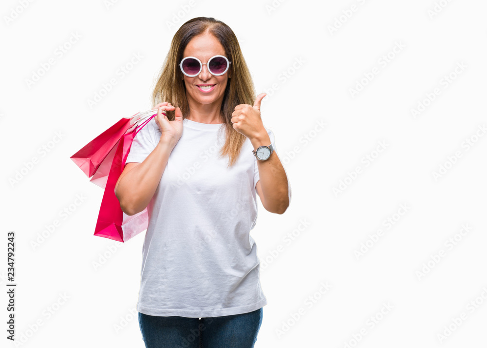 Middle age hispanic woman holding shopping bags on sales over isolated background happy with big smile doing ok sign, thumb up with fingers, excellent sign Middle age hispanic woman holding shopping bags on sales over isolated background happy with big smile doing ok sign, thumb up with fingers, excellent sign