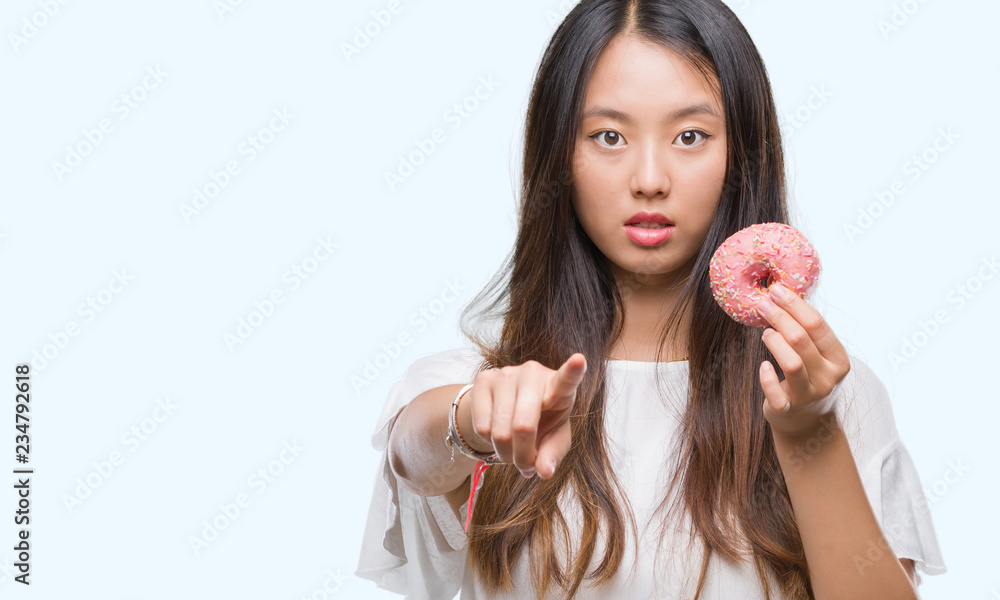 Young asian woman eating donut over isolated background pointing with finger to the camera and to you, hand sign, positive and confident gesture from the front Young asian woman eating donut over isolated background pointing with finger to the camera and to you, hand sign, positive and confident gesture from the front