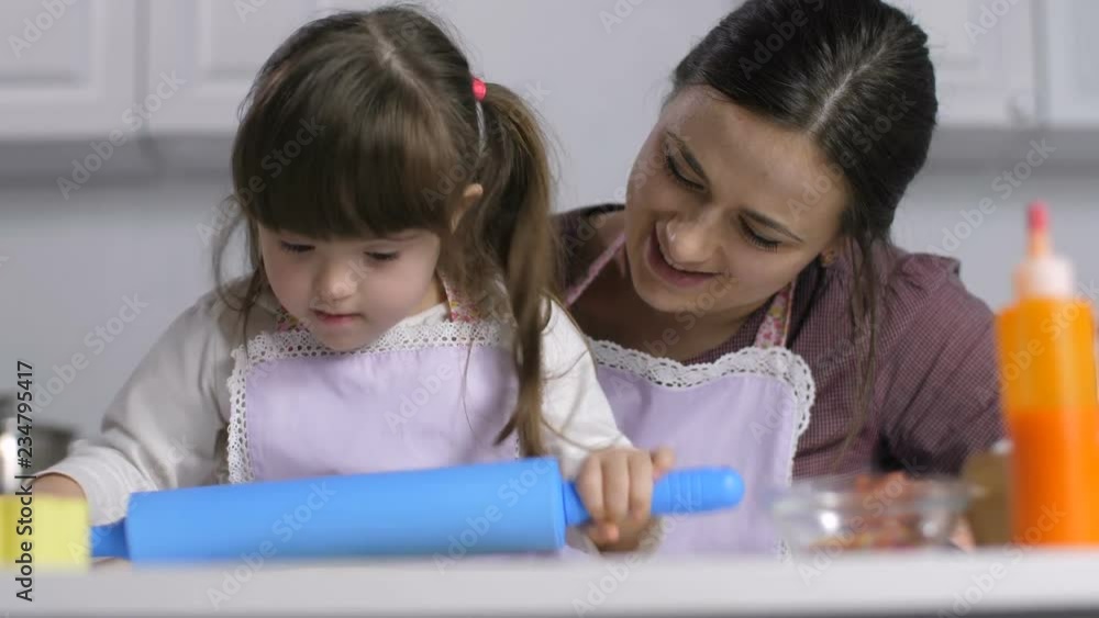 Portrait of concentrated little girl with down syndrome rolling out dough with rolling pin on the kitchen table while mother teaching child to cook. Family with special needs child cooking together. Portrait of concentrated little girl with down syndrome rolling out dough with rolling pin on the kitchen table while mother teaching child to cook. Family with special needs child cooking together.