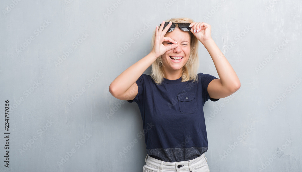 Adult caucasian woman over grunge grey wall wearing sunglasses doing ok gesture with hand smiling, eye looking through fingers with happy face.
