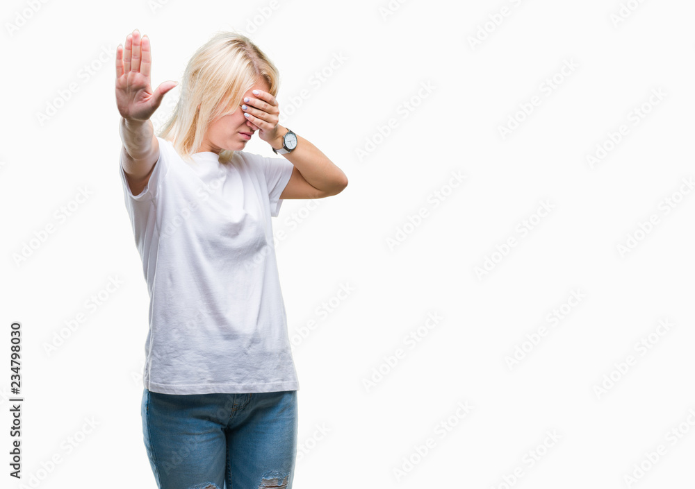 Young beautiful blonde woman wearing white t-shirt over isolated background covering eyes with hands and doing stop gesture with sad and fear expression. Embarrassed and negative concept. Young beautiful blonde woman wearing white t-shirt over isolated background covering eyes with hands and doing stop gesture with sad and fear expression. Embarrassed and negative concept.