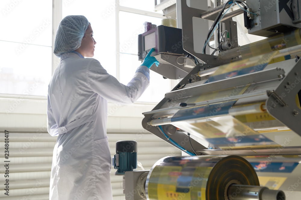 Side view portrait of female factory worker wearing clean white coat standing by machine units at packaging line, copy space