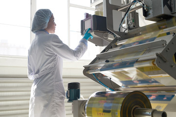 Side view portrait of female factory worker wearing clean white coat standing by machine units at packaging line, copy space