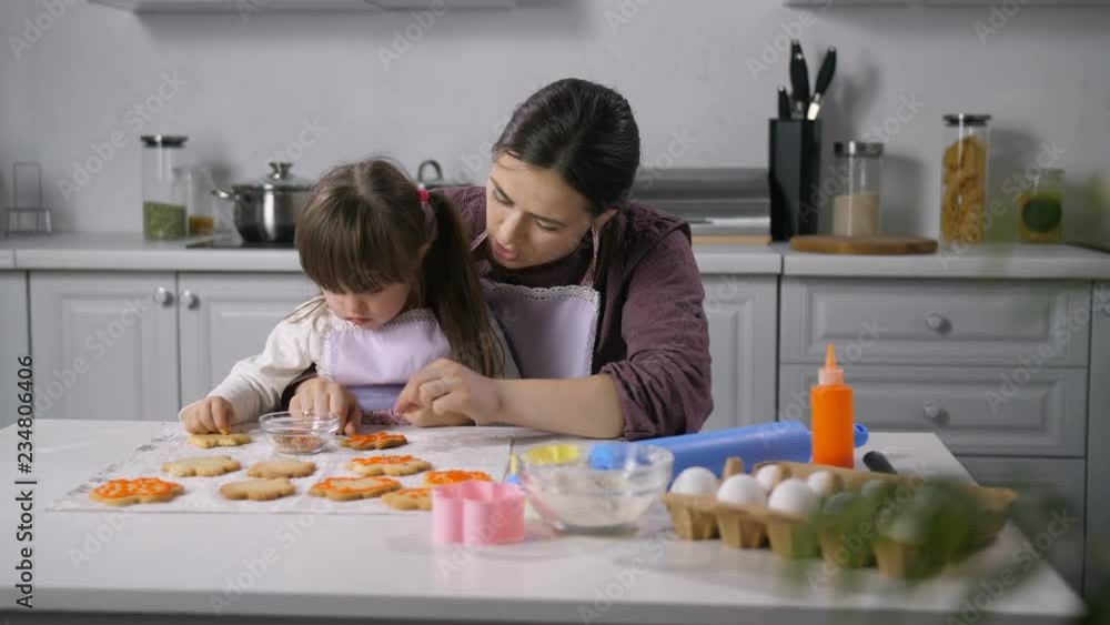 Positive little girl with down syndrome helping happy mother to decor freshly baked biscuits with royal icing in the kitchen. Caring mother developing child with special needs through teaching to cook Positive little girl with down syndrome helping happy mother to decor freshly baked biscuits with royal icing in the kitchen. Caring mother developing child with special needs through teaching to cook