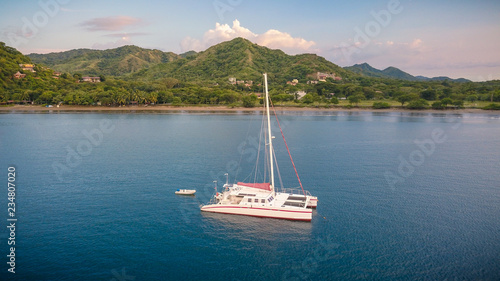A nice drone shot of a large white catamaran yacht anchoring in a beautiful blue bay in front of an empty beach on the green coast of Costa Rica in Central America.