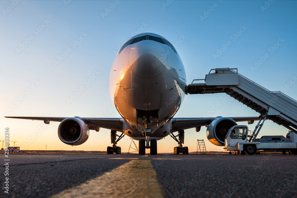 Fototapeta premium Widok z przodu samolotu pasażerskiego białego szerokokadłubowego ze schodami na pokład na płycie lotniska w wieczornym słońcu