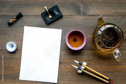Lesson of calligraphy concept. White blank sheet for lettering near writing accessories and tea on dark wooden background top view mockup