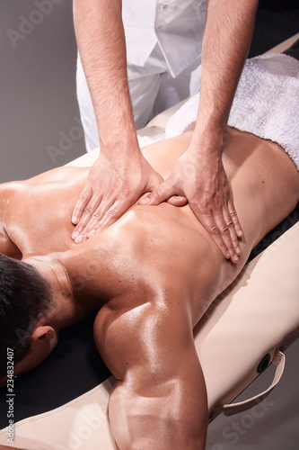 two young man, 20-29 years old, sports physiotherapy indoors in studio, photo shoot. Physiotherapist massaging muscular patient shoulder with his hands close-up.