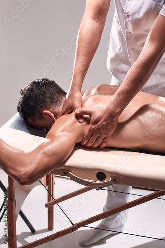 side view, two young man, 20-29 years old, sports physiotherapy indoors in studio, photo shoot. Physiotherapist massaging patient shoulder with his hands.