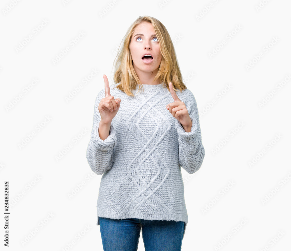Beautiful young woman wearing winter sweater over isolated background amazed and surprised looking up and pointing with fingers and raised arms. Beautiful young woman wearing winter sweater over isolated background amazed and surprised looking up and pointing with fingers and raised arms.