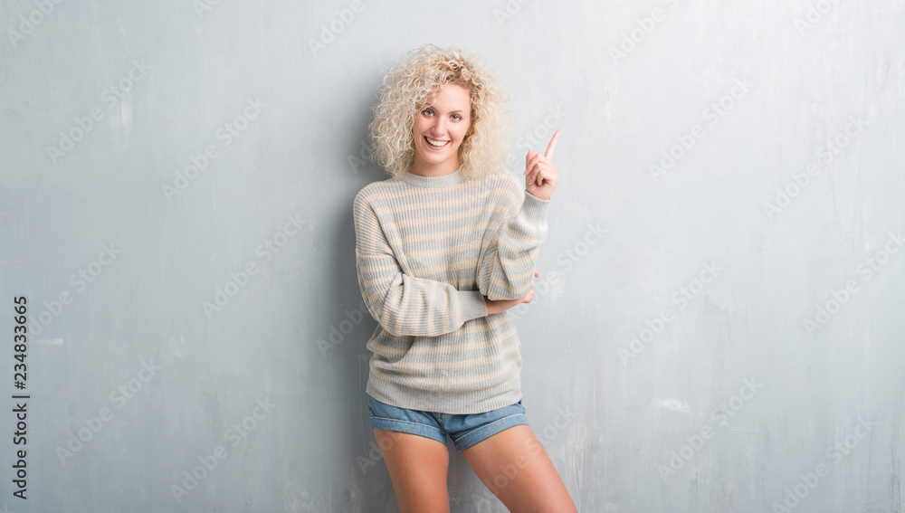 Young blonde woman with curly hair over grunge grey background with a big smile on face, pointing with hand and finger to the side looking at the camera. Young blonde woman with curly hair over grunge grey background with a big smile on face, pointing with hand and finger to the side looking at the camera.
