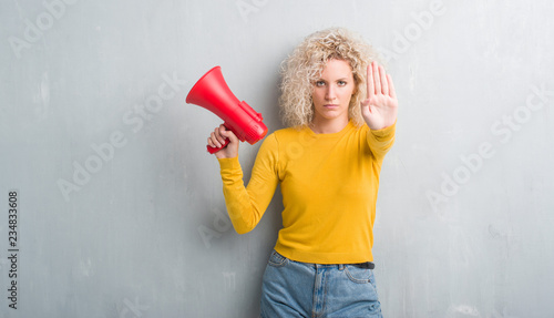 Young blonde woman over grunge grey background holding megaphone with open hand doing stop sign with serious and confident expression, defense gesture
