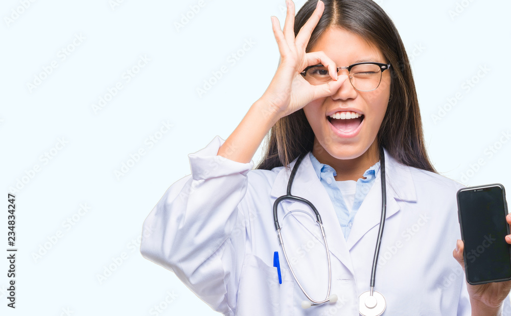 Young asian doctor woman holding smartphone over isolated background with happy face smiling doing ok sign with hand on eye looking through fingers Young asian doctor woman holding smartphone over isolated background with happy face smiling doing ok sign with hand on eye looking through fingers