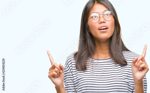 Young asian woman wearing glasses over isolated background amazed and surprised looking up and pointing with fingers and raised arms.