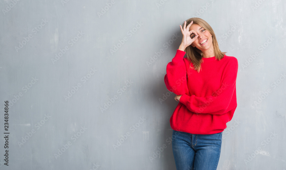 Beautiful young woman standing over grunge grey wall wearing winter sweater with happy face smiling doing ok sign with hand on eye looking through fingers Beautiful young woman standing over grunge grey wall wearing winter sweater with happy face smiling doing ok sign with hand on eye looking through fingers