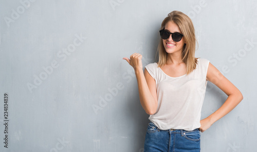 Beautiful young woman standing over grunge grey wall wearing fashion sunglasses pointing and showing with thumb up to the side with happy face smiling