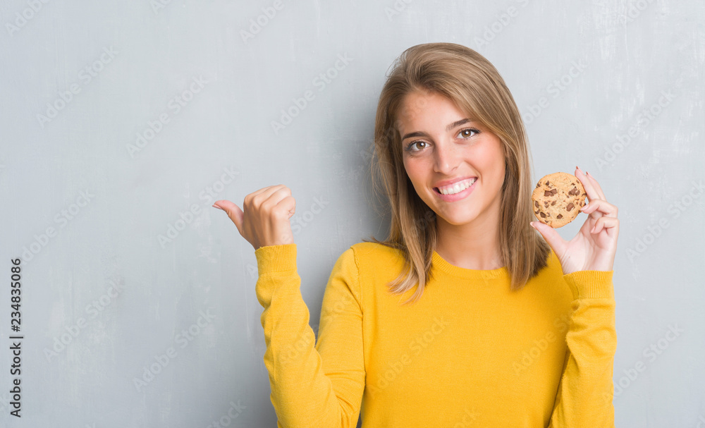 Beautiful young woman over grunge grey wall eating chocolate chip cooky pointing and showing with thumb up to the side with happy face smiling Beautiful young woman over grunge grey wall eating chocolate chip cooky pointing and showing with thumb up to the side with happy face smiling