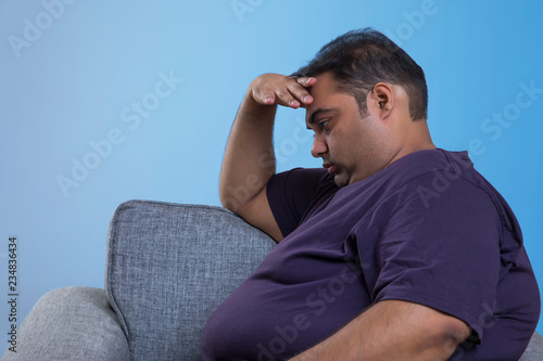 Side view of worried obese man sitting on sofa with hand on forehead